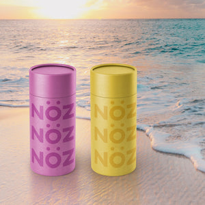 Fluorescent Prurple and Bright Yellow Noz sunscreen on the sand in front of the ocean sunset